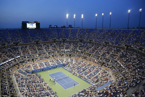 Stadio pieno agli US Open di tennis con una partita serale in corso all'Arthur Ashe Stadium di New York, che mostra il grande pubblico e il campo illuminato.