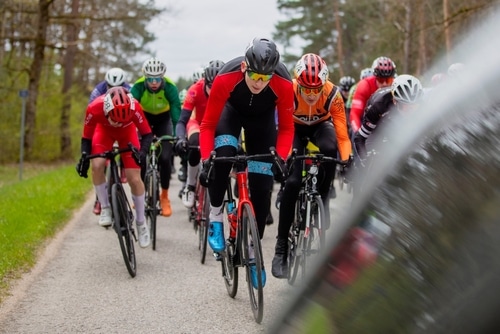 Gruppe von Radfahrern, die an einem Straßenrennen teilnehmen, mit dem führenden Radfahrer in einem rot-schwarzen Outfit, umgeben von anderen Radfahrern in bunten Trikots, auf einer von Bäumen gesäumten Landstraße.