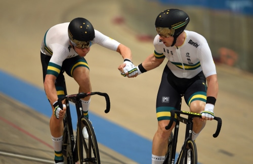 Two Australian cyclists in white and green racing uniforms, holding hands during a track cycling event, demonstrating teamwork and camaraderie while riding their bikes on an indoor velodrome.