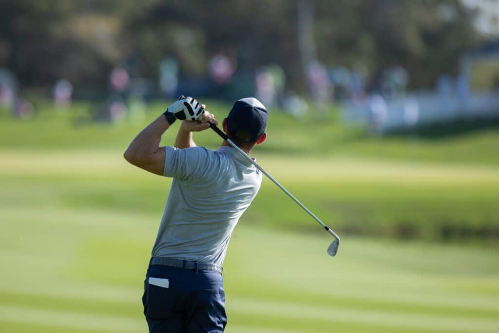 A golfer in a grey shirt and navy cap swings a golf club on a green course, focusing on a powerful shot, with blurred background featuring spectators in the distance.